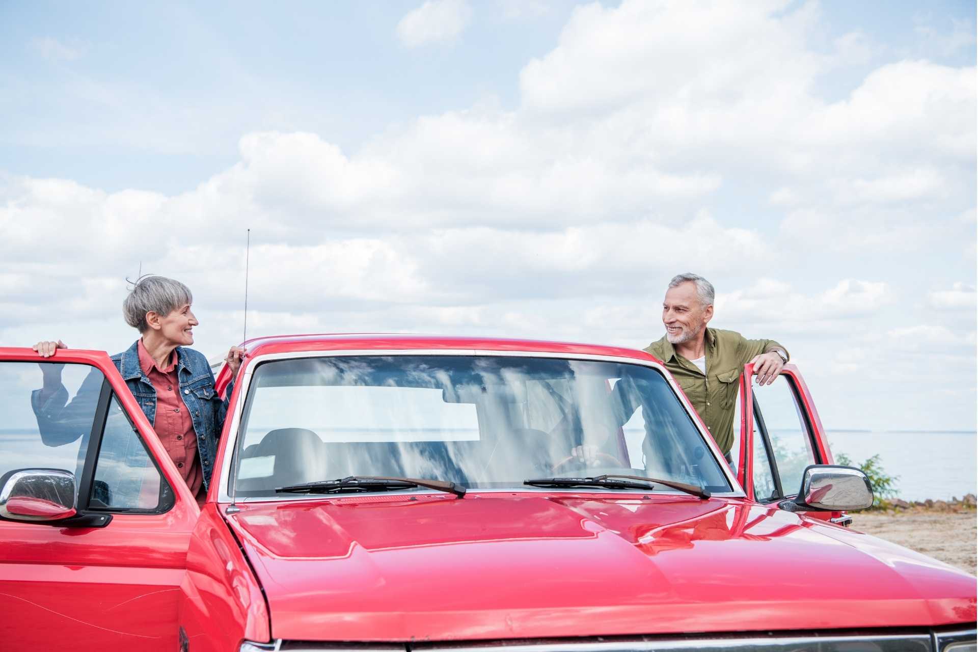 A senior couple smiling at each other as they get into a red vintage car.